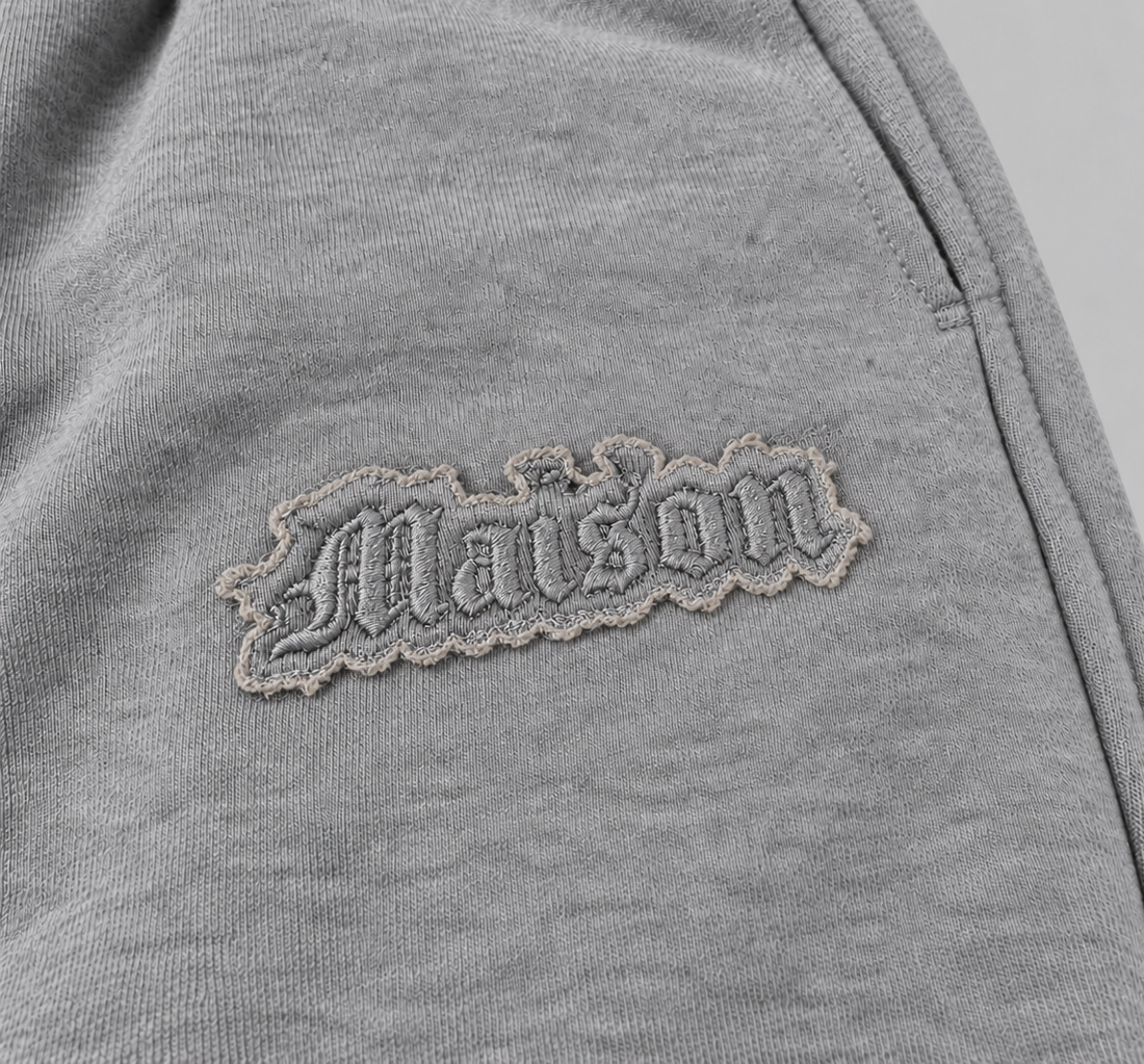 Signature Sweatpants Product V3 Grey Melange. Nahaufnahme des gestickten Logos auf den grauen Maison De La Rue Sweatpants.